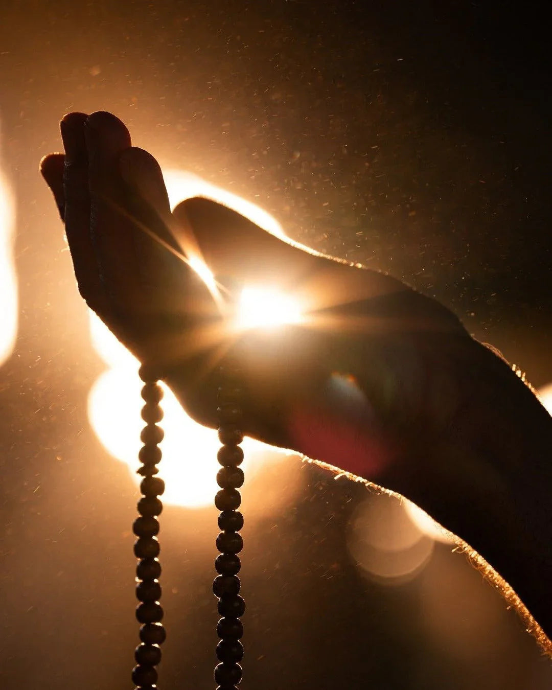 Hand holding wooden prayer beads with warm sunlight in background and dust particles visible