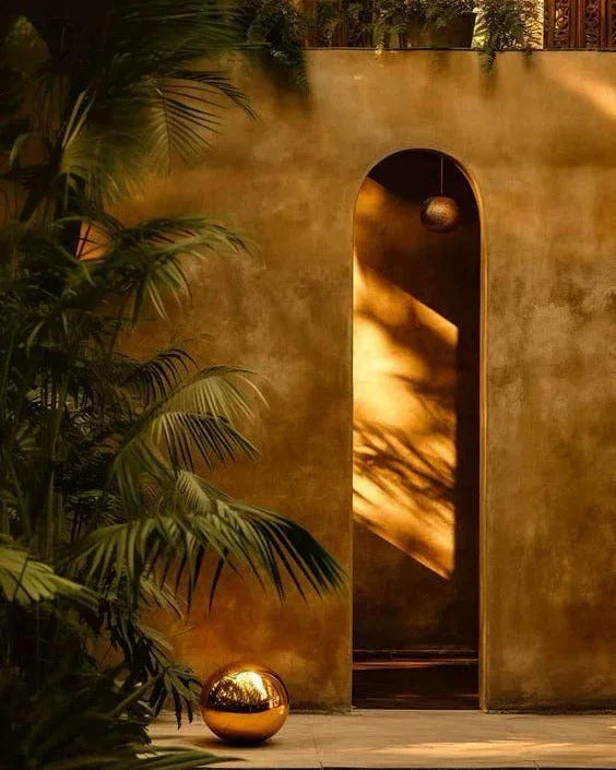 Tropical green plants beside a textured golden wall with arched doorway and reflective golden orb on floor